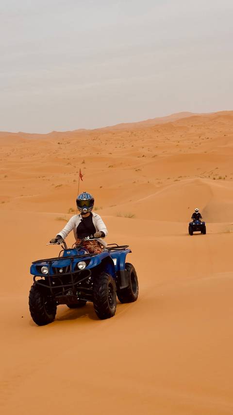       Two riders on blue quad bikes navigating rolling desert sand dunes.
  