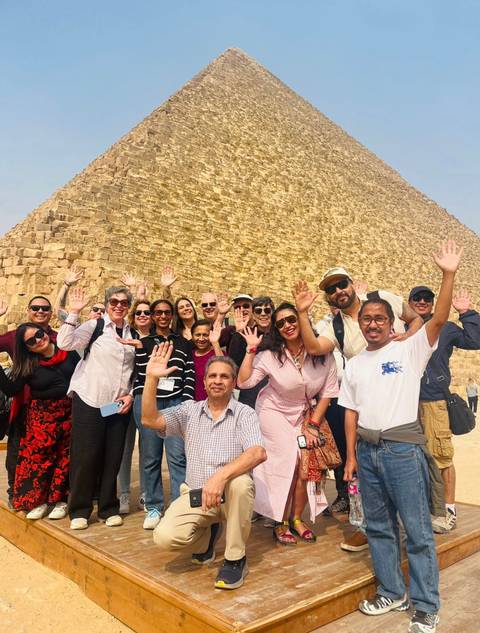       Happy tour group posing with raised hands in front of the Great Pyramid’s massive stone blocks
  