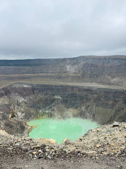       Steaming crater of Santa Ana volcano with layered rock walls and pale green acidic lake at bottom.
  