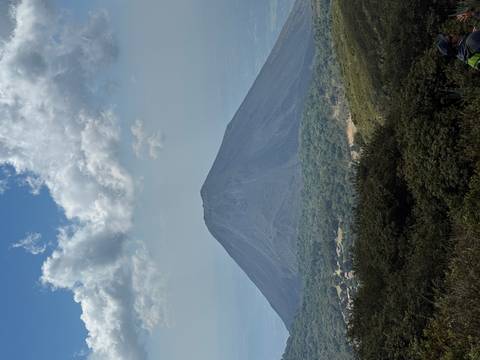       Conical volcano rising above forested foothills under partly cloudy sky.
  