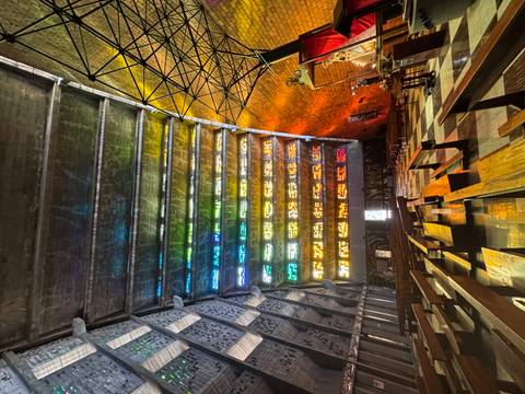       Interior of modern church with colourful stained-glass light strips illuminating wooden pews.
  
