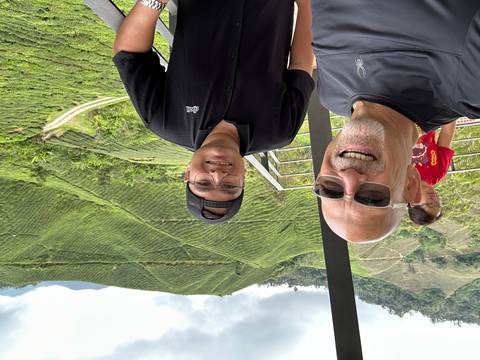       Two men smiling for a selfie on a viewing deck overlooking the lush tea plantations of Cameron Highlands.
  