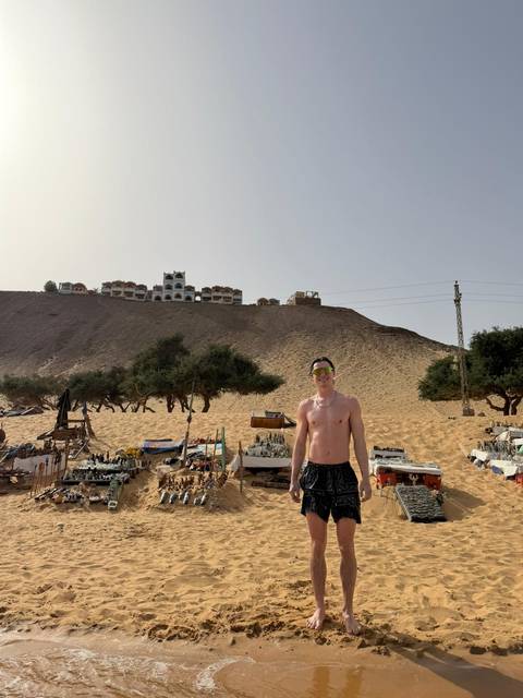       Shirtless traveler stands before a sandy hillside market on the edge of the desert.
  