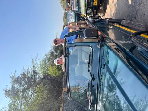       A couple smiles from the open roof of a dark green safari jeep ready for a wildlife drive.
  