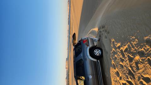       4x4 vehicle parked on rippling golden desert dunes at dawn with distant cliffs on the horizon.
  