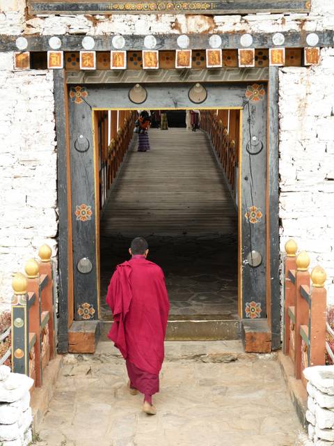       Young monk in red robes walking across a wooden covered bridge into a Bhutanese dzong
  