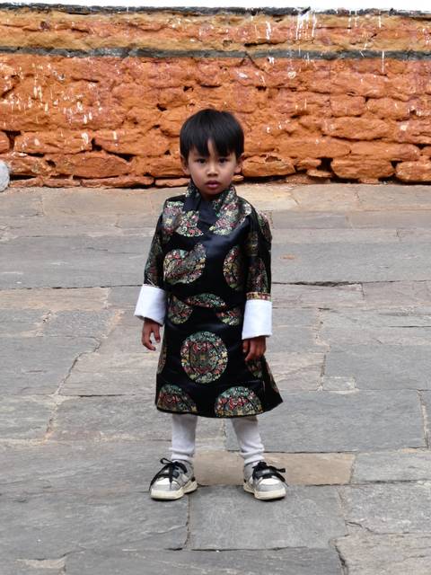       Young Bhutanese boy in an elegant traditional robe standing on a stone courtyard.
  