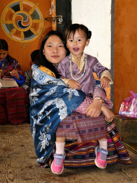       Woman in traditional dress holding a child adorned with beads inside a Bhutanese interior.
  