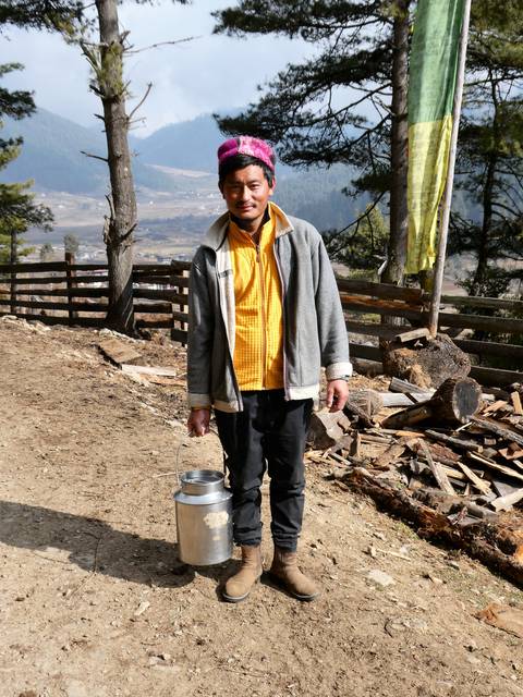       Bhutanese man carrying a metal bucket in a rural mountain setting with firewood piles behind.
  