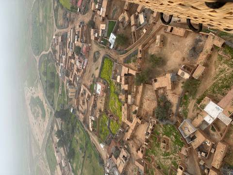       Hazy aerial view of a rural Moroccan village with green crop patches and winding dirt roads.
  