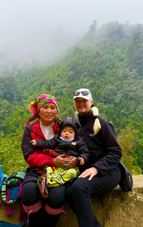       Two women and a baby in colorful ethnic attire stand against a lush mountainous backdrop in Sapa.
  