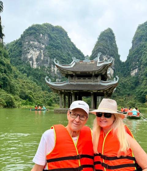       Smiling couple in front of a traditional pavilion amid towering limestone karsts on a tranquil river in Ninh Binh.
  