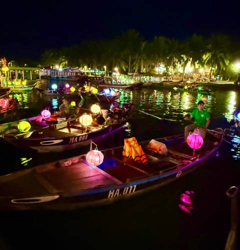       Brightly lit lantern boats float on Hoi An’s river at night, reflections shimmering in the water.
  
