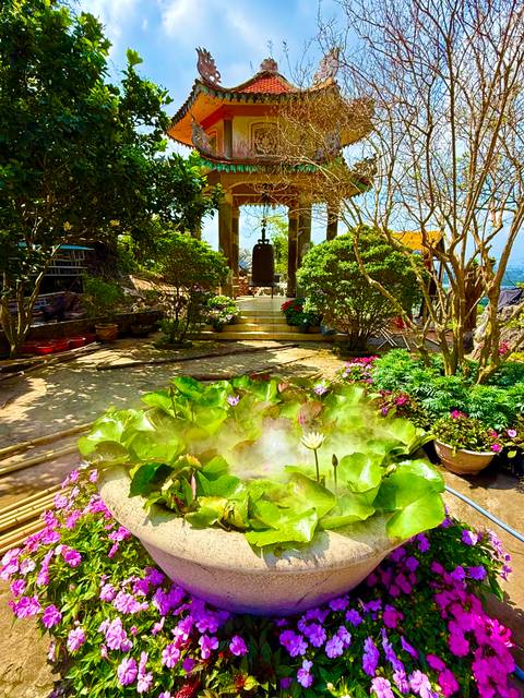       Lush garden courtyard with blooming flowers, lotus pond and a hanging bronze bell in Da Nang’s Marble Mountains area.
  
