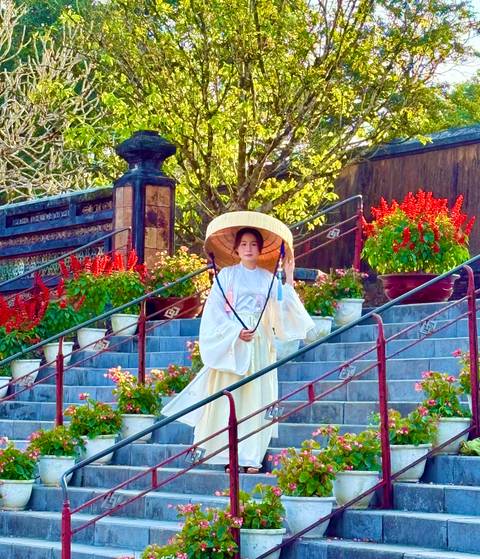       Woman in traditional white ao dai and conical hat stands gracefully on stone steps amid red flowers in Hue.
  