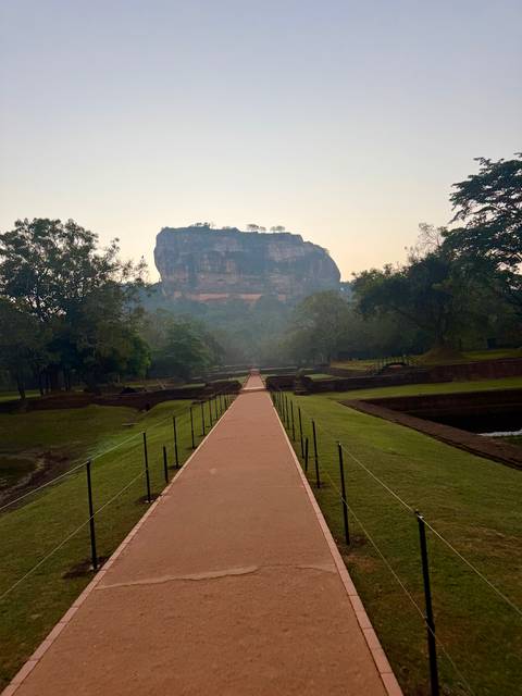       Straight walkway leading toward the towering Sigiriya Rock surrounded by green gardens in soft morning haze.
  
