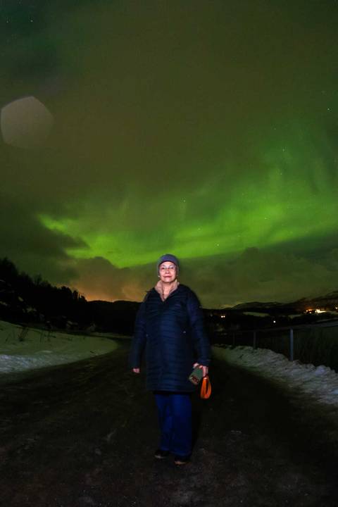       Portrait of a traveler standing under vivid green Northern Lights with snowy hills in Tromsø.
  