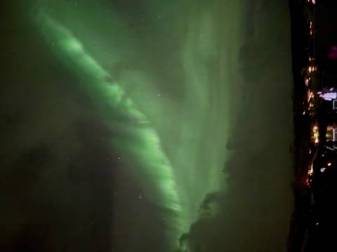       Vertical display of Northern Lights arcing over faintly lit buildings at night.
  