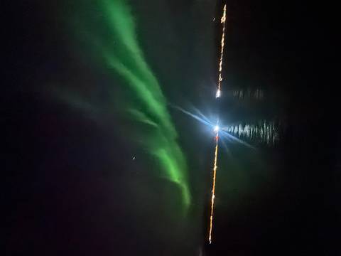       A sweeping green aurora arcs above calm dark water and distant town lights, pierced by a bright upward beam.
  