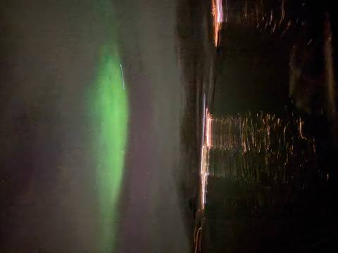       Dim green aurora glow hovers over blurred waterfront lights reflected in the dark water.
  