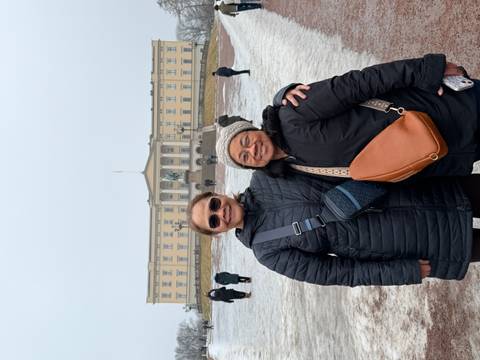       Two women in winter coats stand smiling before a yellow palace building across a snowy square.
  