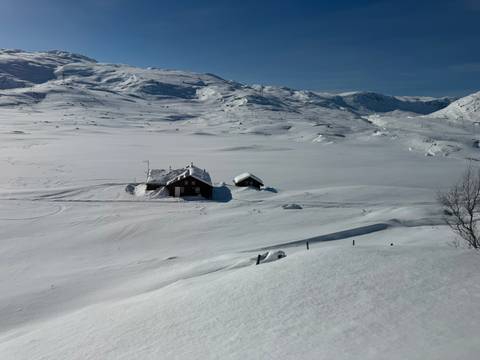       Remote wooden cabins sit within a vast snow-covered mountain plateau under a clear blue sky.
  