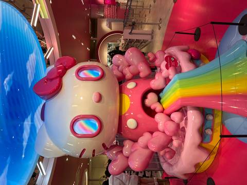       Giant pink cartoon cat statue with rainbow slide stands inside a colourful themed store.
  