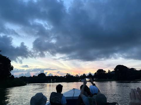       Dramatic sunset colours spread across cloudy skies above a reflective rainforest river as travellers photograph the view.
  