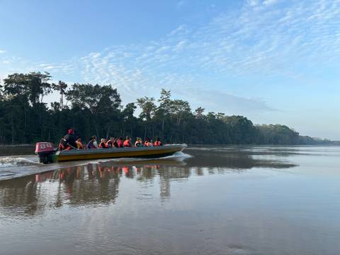       Longboat with life-jacketed tourists speeds along calm rainforest river at dawn.
  