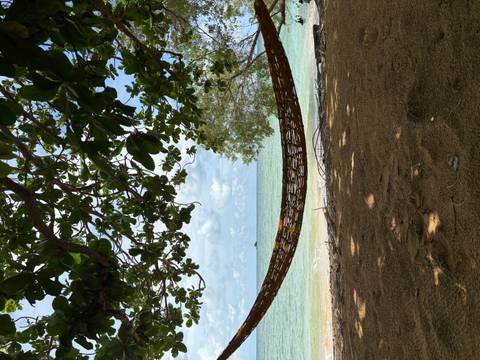       Woven hammock strung under leafy shade facing turquoise sea and distant islet.
  