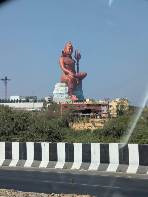       Gigantic seated statue of a Hindu deity towers above surrounding buildings under a clear blue sky.
  