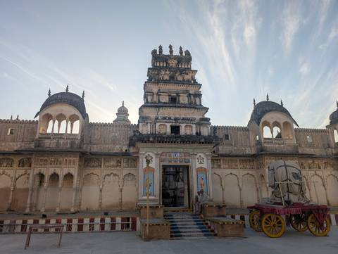       Historic Indian temple complex with arched gateways, faded frescoes and three domes under streaked morning sky.
  