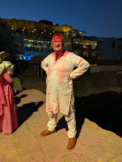      Man wearing a kurta stained with Holi colors stands in a dimly lit courtyard at night.
  
