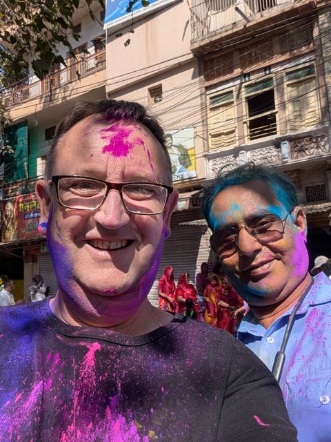       Close selfie of two men covered in bright Holi powders smiling in a busy street.
  
