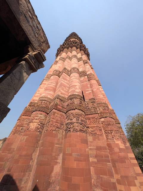       Low-angle view of the red-sandstone Qutub Minar tower with intricate carvings against clear sky.
  