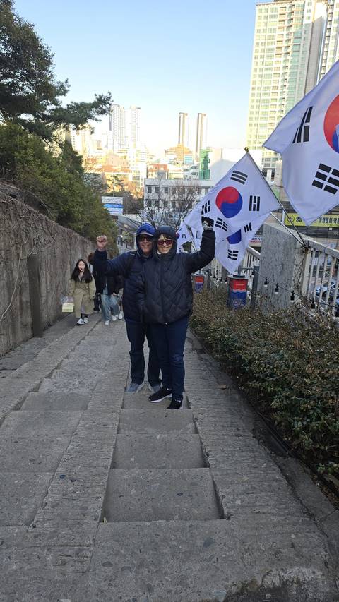       Smiling travellers raise fists beside South Korean flags on a stone stair pathway.
  
