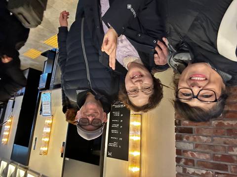       Tourists and local woman smile for selfie inside a warm-lit indoor space.
  