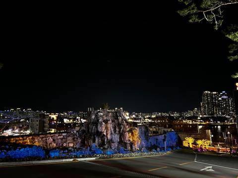       Night city skyline behind illuminated rocky sculpture with lights twinkling across horizon.
  