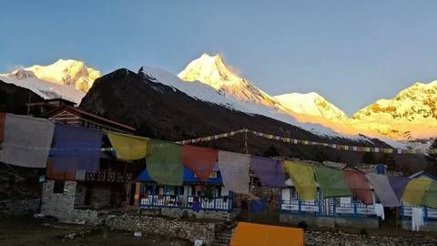       Colorful prayer flags flutter outside a small Himalayan village with sunrise light on snowy peaks.
  