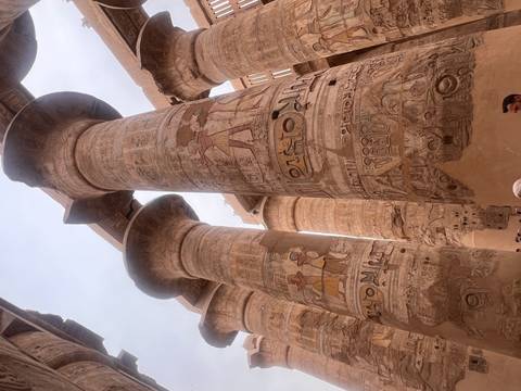       Massive decorated stone columns of Karnak Temple with faint paintings and visitors dwarfed below.
  