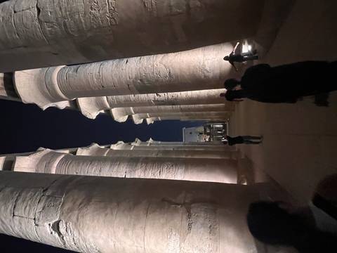       Nighttime corridor of illuminated stone columns at Luxor Temple with visitors walking through.
  
