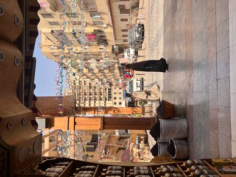       Daytime street scene in an Egyptian city with two locals in traditional clothing walking past market stalls and apartment blocks decorated with colorful streamers.
  
