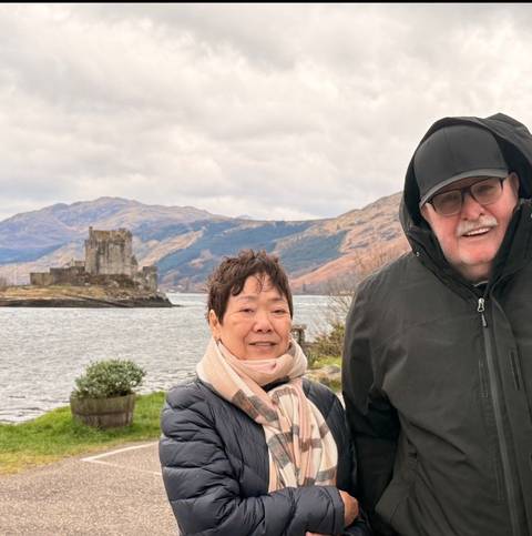       Two older travellers in winter jackets pose in front of a historic stone castle on a small loch island with rolling Scottish hills behind them under a cloudy sky.
  