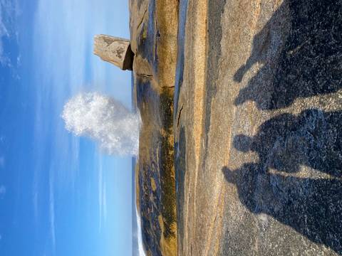       Sea spray erupts from a coastal blowhole as three travellers’ shadows stretch across sun-lit granite.
  