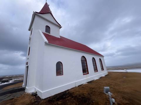       A small white church with a red roof and arched windows stands alone on a windswept grassy ridge beneath a brooding grey sky.
  