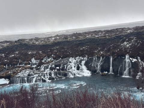       Blue-tinged water cascades in countless icy rivulets over dark basalt ledges at Hraunfossar waterfall on a grey winter day.
  