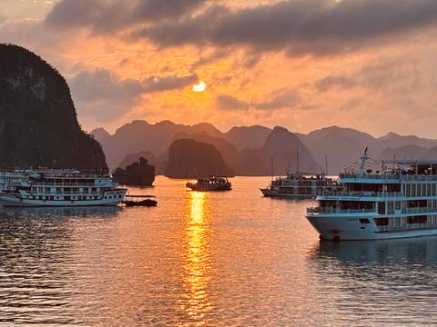       Orange sunset over Halong Bay with cruise boats reflecting on calm water and limestone karsts in the distance.
  