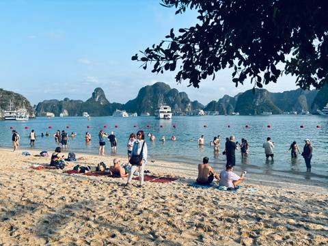       Tourists swim and relax on a sandy beach with Halong Bay’s limestone islands in the background.
  