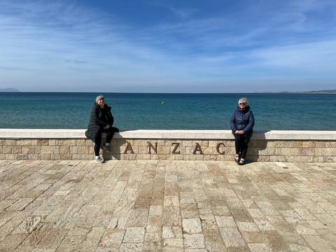      Two women sit on a stone wall marked ANZAC overlooking the clear blue sea at Gallipoli.
  