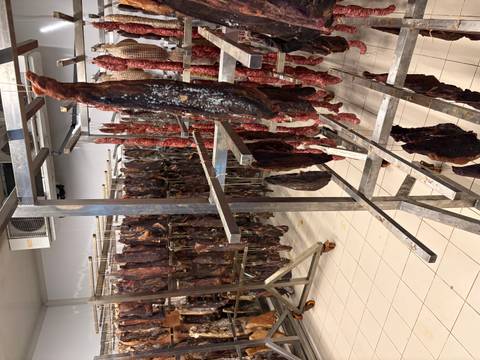       Rows of cured meat and sausages hanging on metal racks inside a processing room.
  